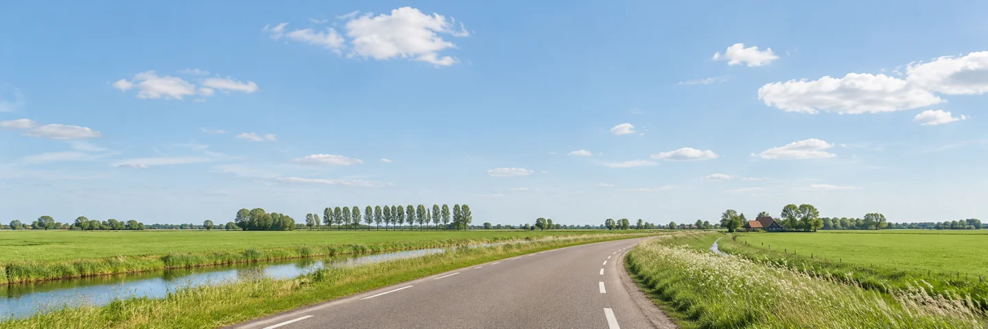 Een lege polderweg tussen weilanden en een sloot, onder een blauwe lucht.