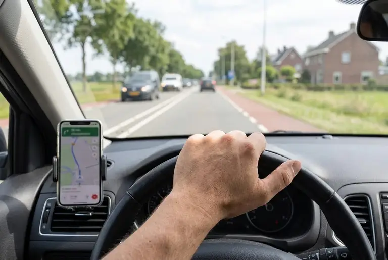 Een telefoon met een kaart in een houder op het dashboard, de bestuurder rijdt over een Nederlandse weg.